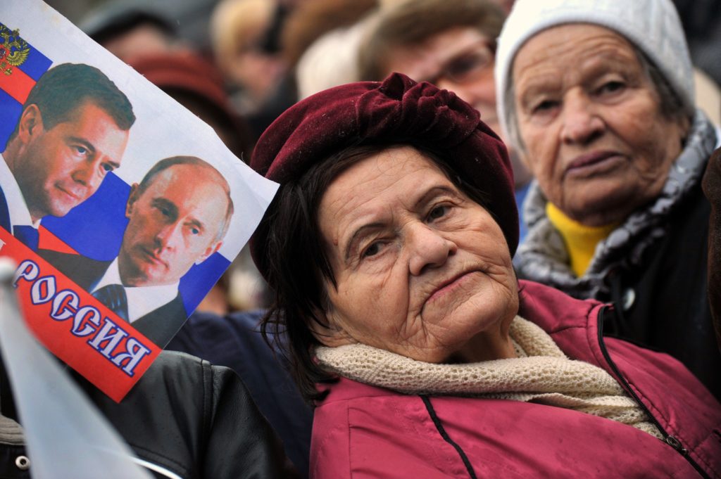 An elderly woman stands next to a flag depicting Russian President Vladimir